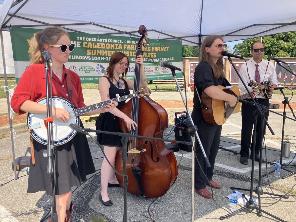 Four musicians perform outdoors under a tent, playing banjo, upright bass, guitar, and standing microphone. A banner behind them promotes a farmers market summer music series.