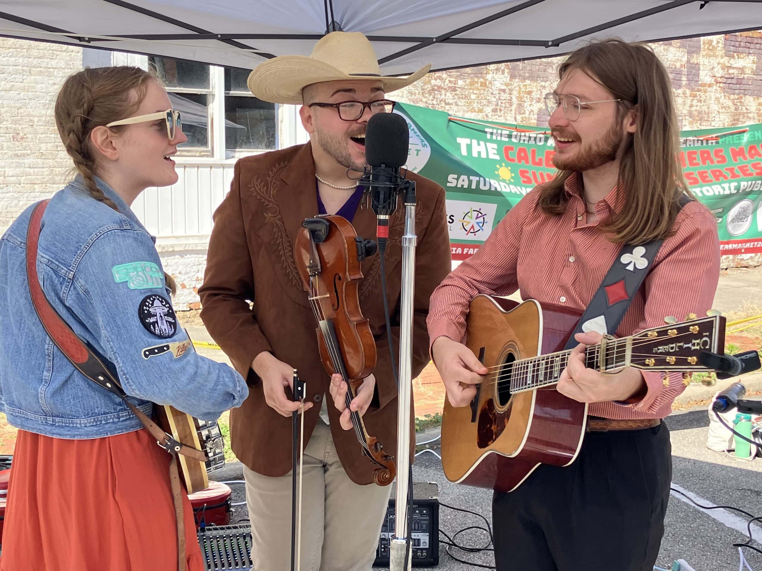 Three musicians stand under a canopy, singing and playing instruments including a guitar, fiddle, and banjo at an outdoor event.