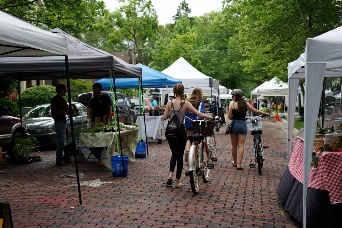 Three people walk bicycles through an outdoor market with vendor tents lining both sides of a brick-paved street on a cloudy day.