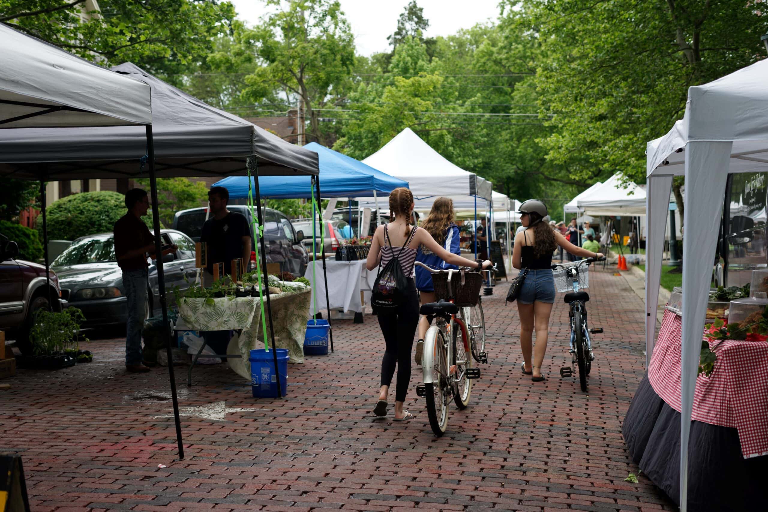 Three people walk bicycles through an outdoor market with vendor tents lining both sides of a brick-paved street on a cloudy day.