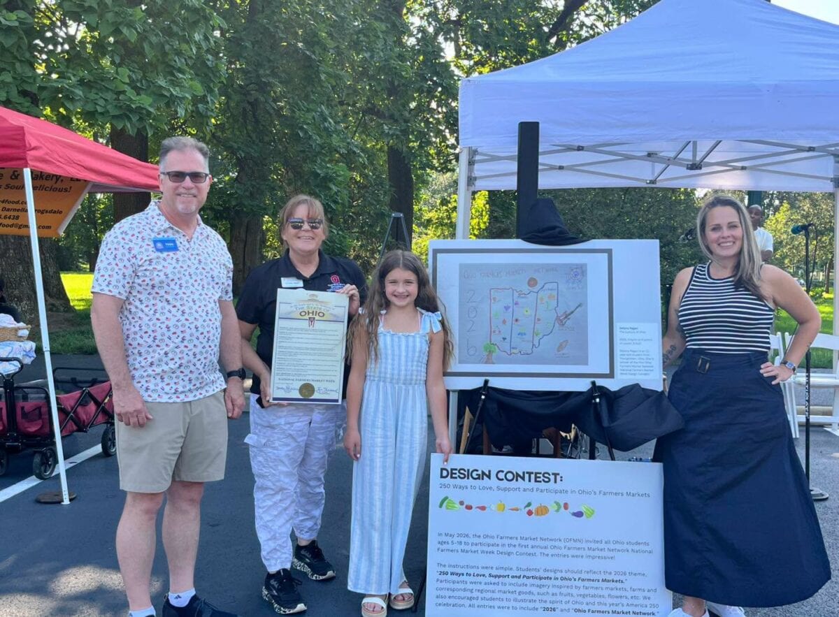 Four people stand outdoors beside a display featuring a design contest poster and a large illustrated map, with trees and tents visible in the background.