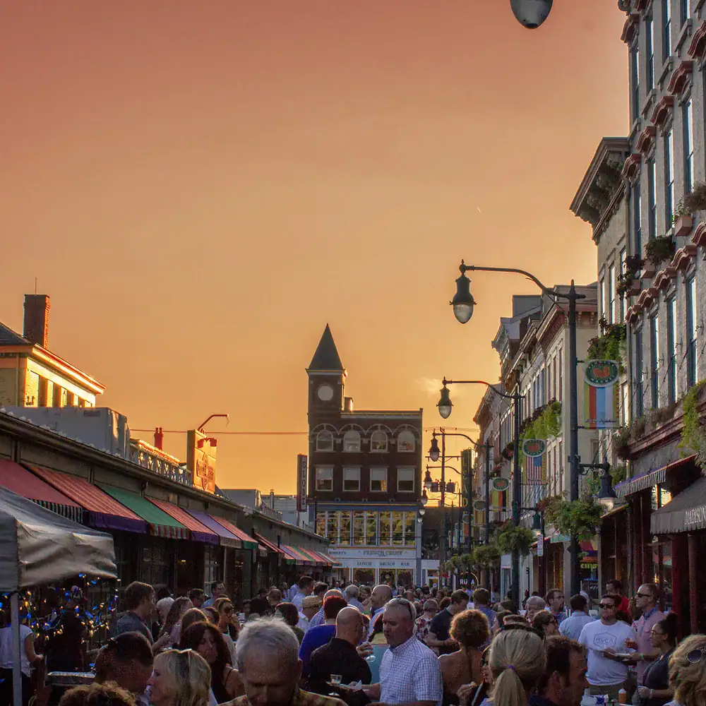 A large crowd fills a city street lined with buildings at sunset, with a clock tower visible in the background.