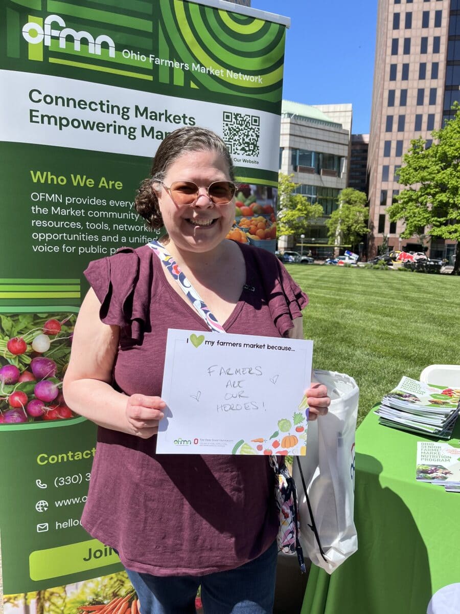 A woman stands outdoors holding a sign that says “Farmers are our heroes” at an Ohio Farmers Market Network booth with informational banners and brochures.