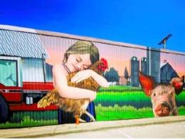 Mural of a young girl hugging a chicken, with a pig, tractor, and farm buildings in the background under a blue sky.