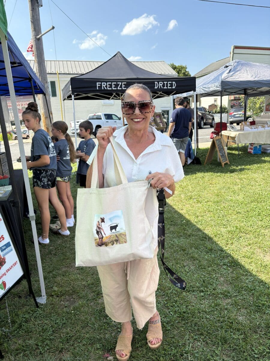 A woman in sunglasses stands on grass at an outdoor market, holding a tote bag with a dog image. Booths and people are visible in the background.