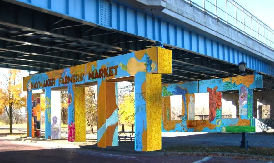 Colorful painted entrance structure for Haymaker Farmers' Market stands beneath a blue bridge, with autumn trees and a brick walkway in the background.
