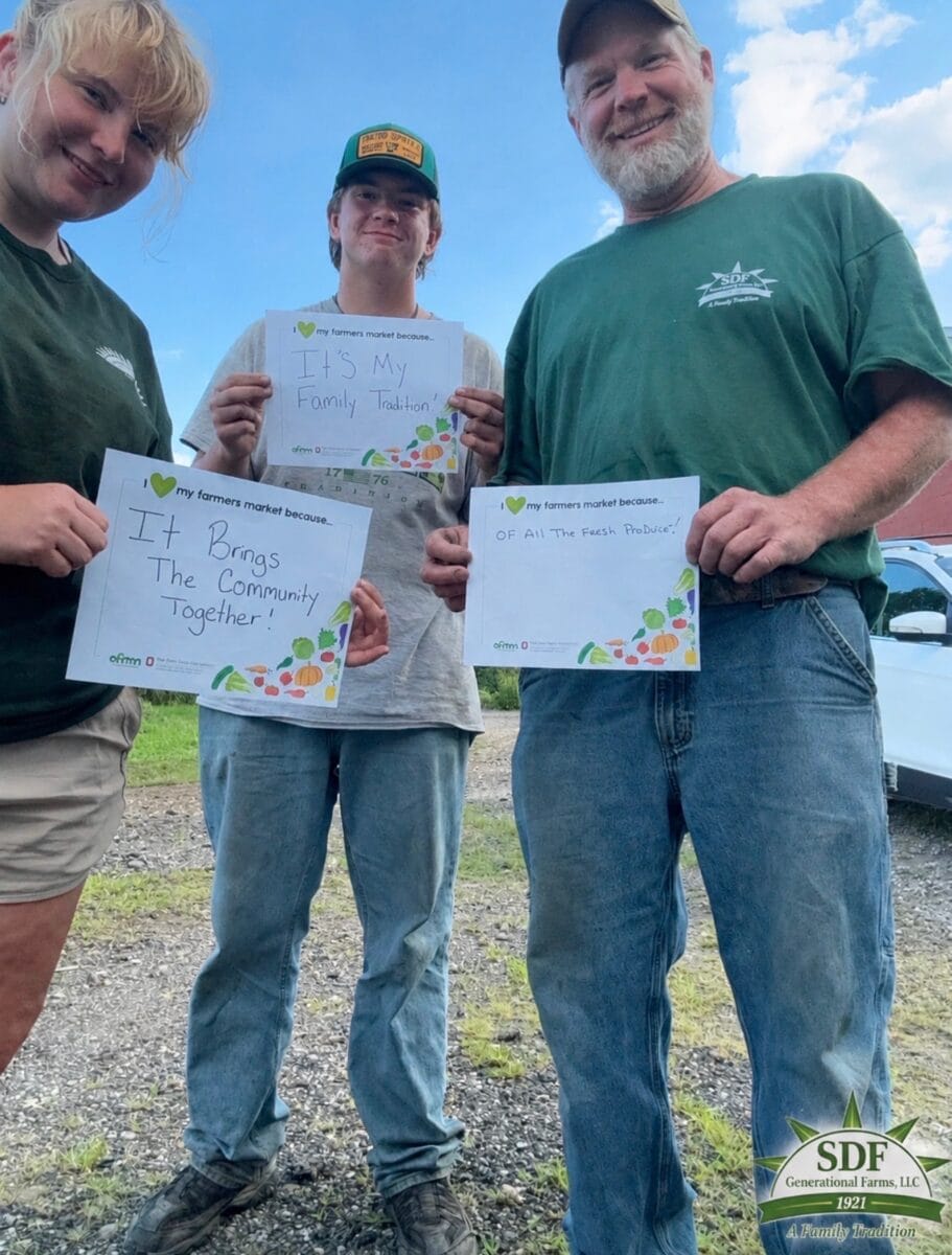 Three people outdoors hold signs about what they love about farmers markets, mentioning family tradition, fresh produce, and community. A logo for Greenfield Farms, LLC is in the bottom corner.