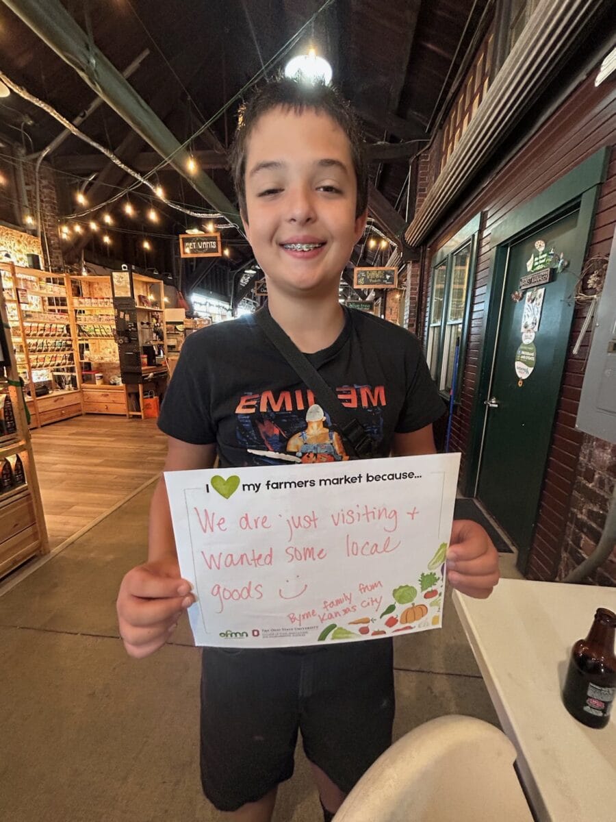 A smiling boy stands indoors holding a sign that says they are visiting the farmers market to get some local goods. Shelves with products and bright lighting are visible in the background.
