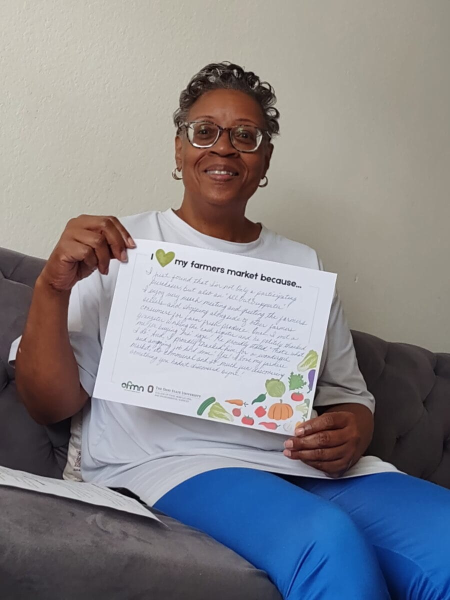 A woman sits on a couch holding a handwritten sign that says “I love my farmers market because...” with text and colorful produce illustrations.