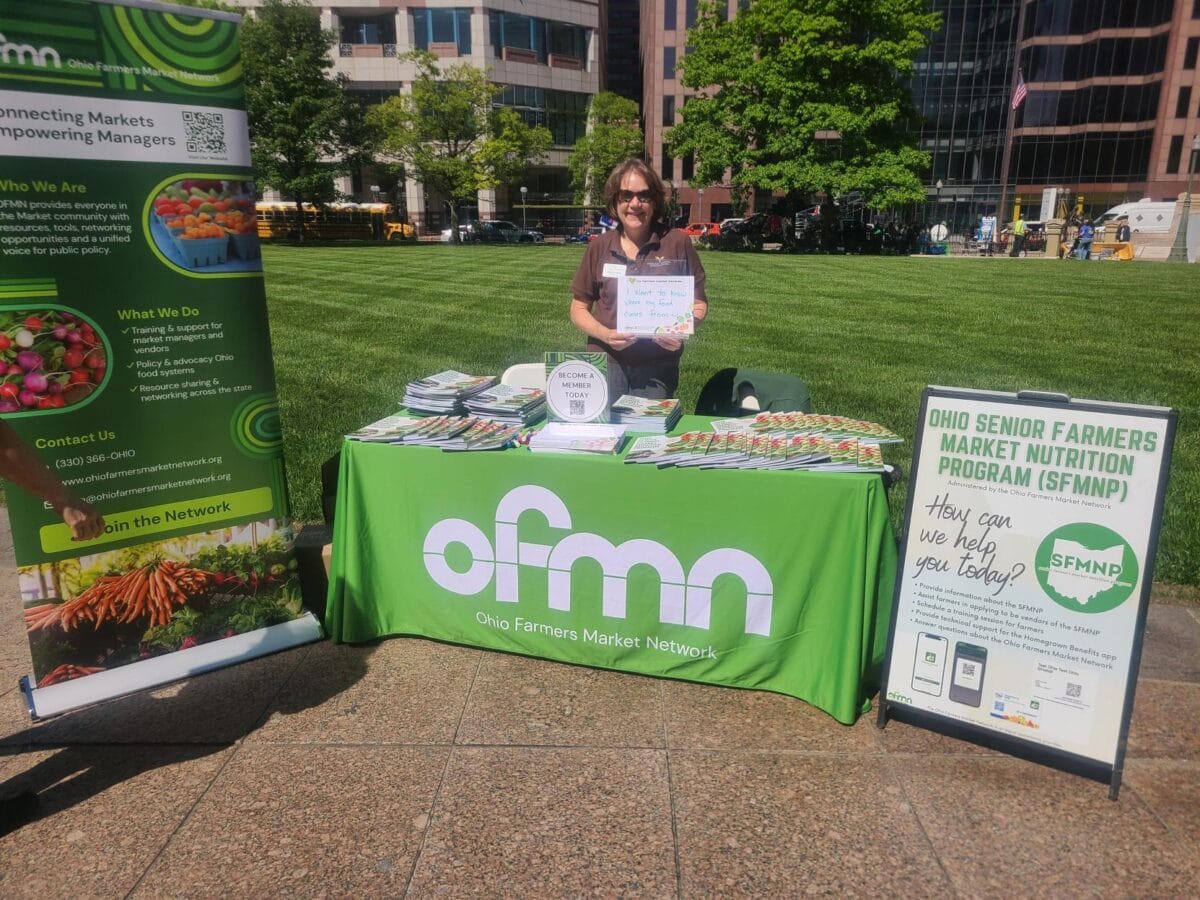 A person stands behind an Ohio Farmers Market Network table with brochures and displays at an outdoor event on a sunny day.