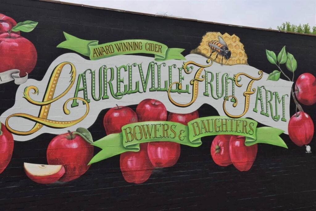 Mural with red apples and green leaves reads "Laurelville Fruit Farm, Award Winning Cider, Bowers & Daughters" with a honeycomb and bee illustration.