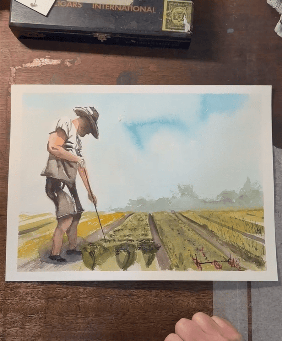 A watercolor painting of a person in a hat tending crops in a field, with blue sky and distant greenery in the background, placed on a wooden table.