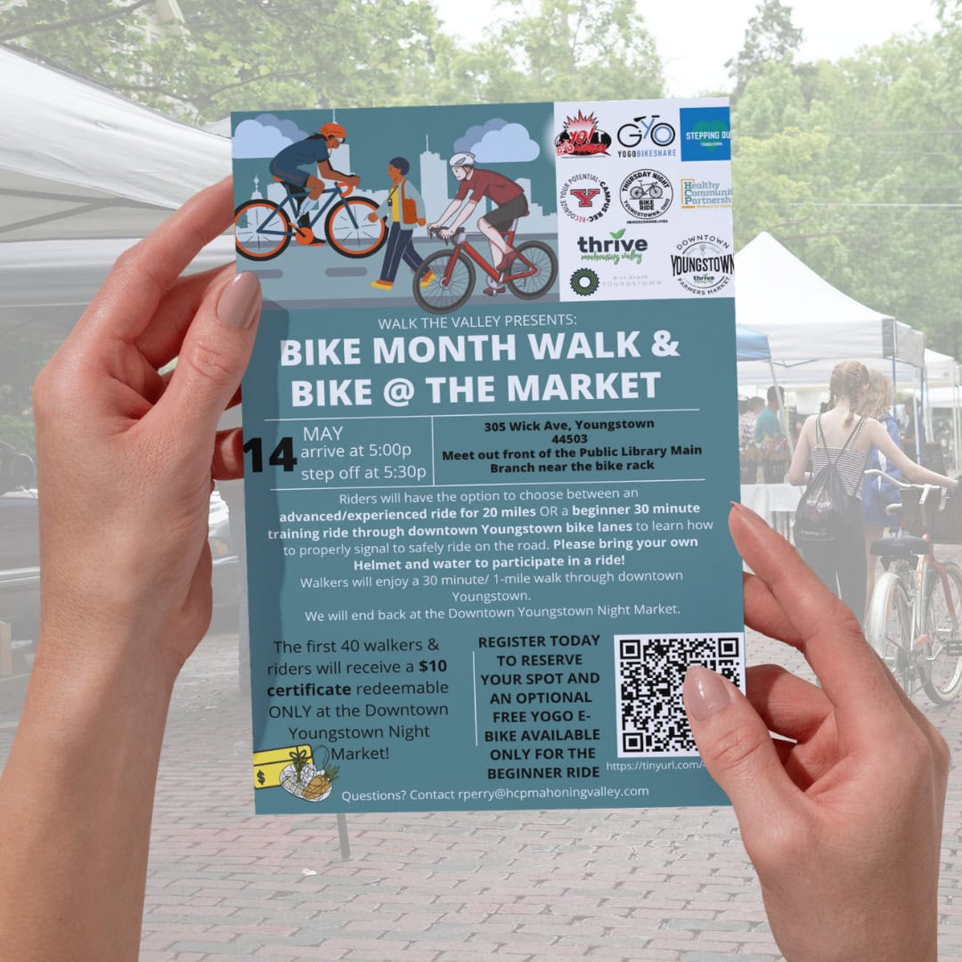 A person holds a flyer promoting the "Bike Month Walk & Bike @ The Market" event in Youngstown, listing details, sponsors, QR code, and contact information, with market tents in the background.