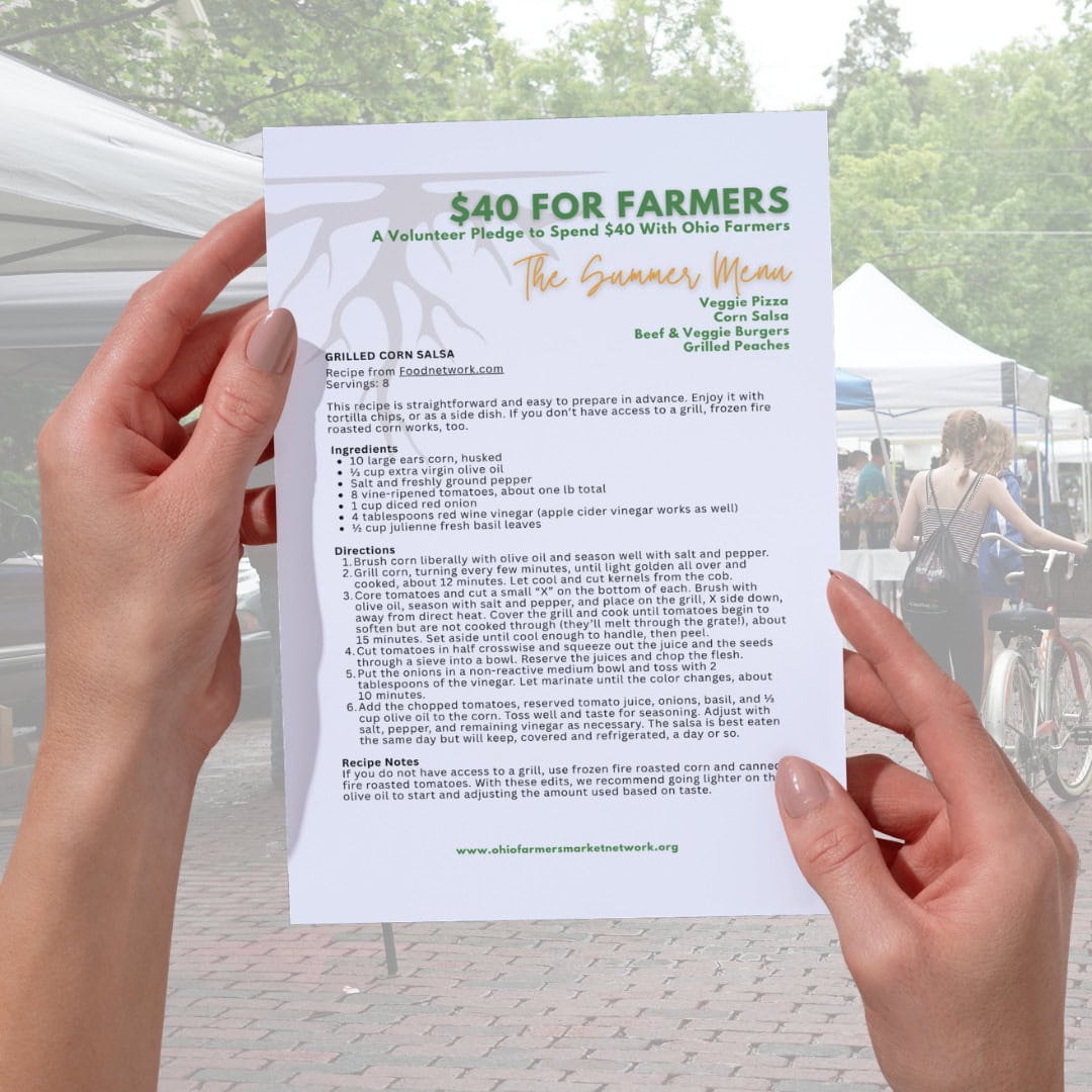 Person holding a printed flyer titled "$40 FOR FARMERS" with a summer menu and recipe details at an outdoor farmers market.