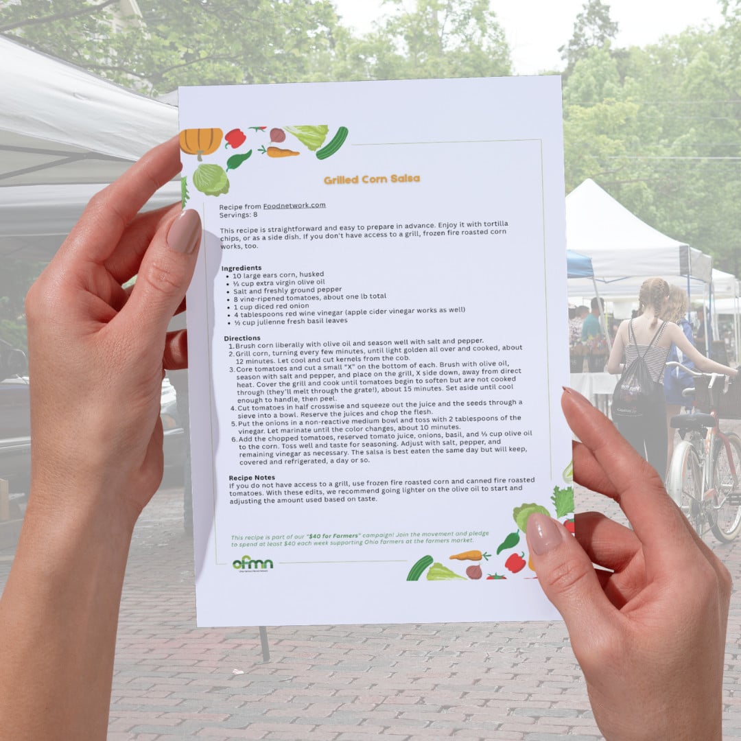 A person holds a printed grilled corn salsa recipe at an outdoor farmers market; market stalls and people are visible in the background.
