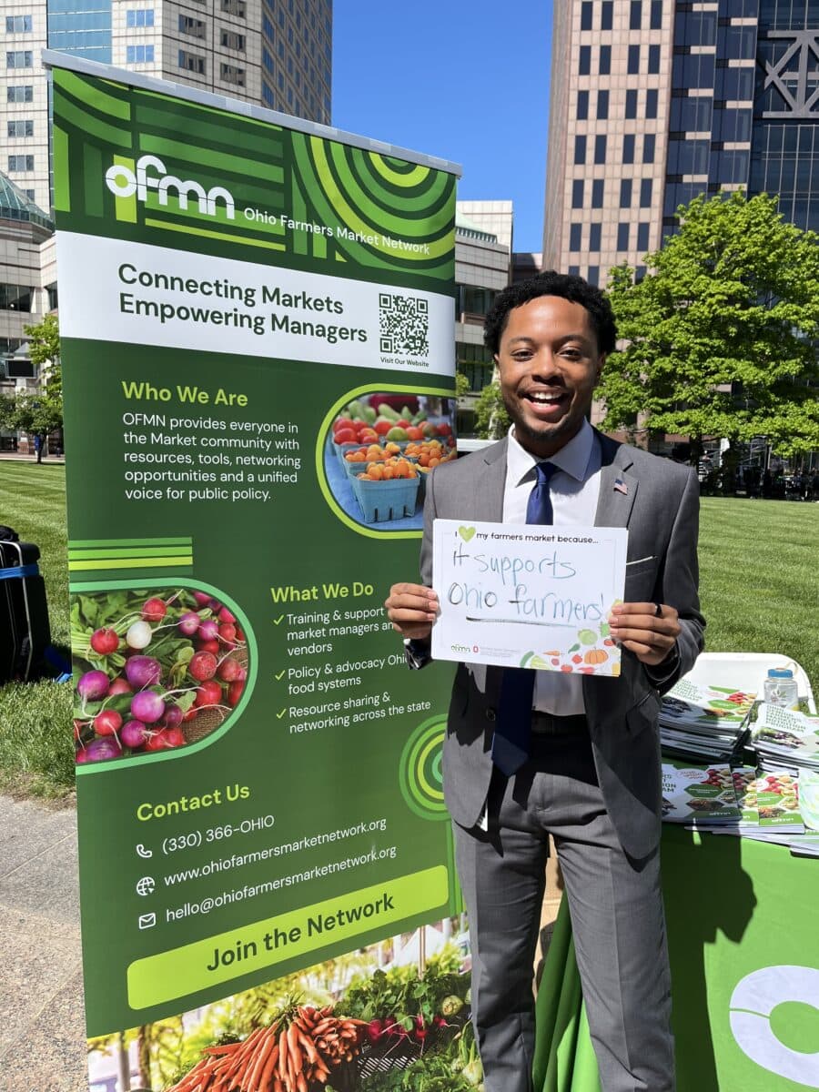 A man in a suit stands outdoors holding a “#supports Ohio farmers” sign in front of an Ohio Farmers Market Network booth with informational banners.