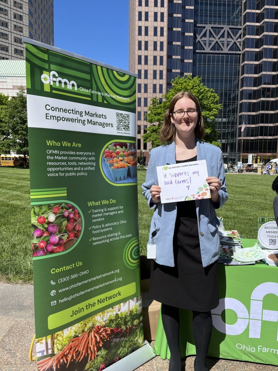 A woman stands outdoors holding a handwritten sign that says, “It supports my local farms!” next to an Ohio Farmers Market Network display booth.
