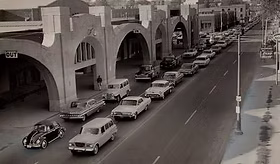 Black and white photo of vintage cars lined up on a city street beside an arched brick building, with pedestrians on the sidewalk.