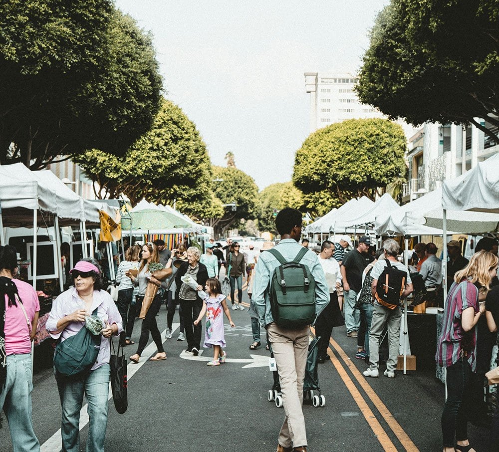 People walking and shopping at an outdoor market with white tents, greenery overhead, and vendors selling goods along a closed street.