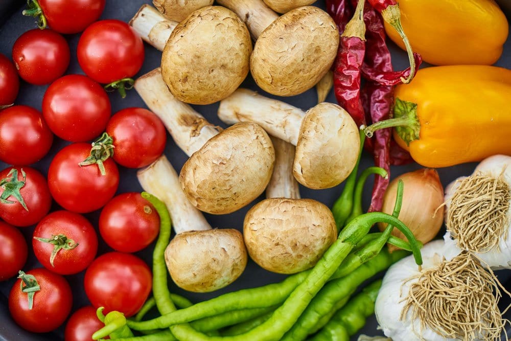 A variety of fresh vegetables including cherry tomatoes, mushrooms, yellow peppers, green chili peppers, garlic, onions, and dried red peppers arranged close together.
